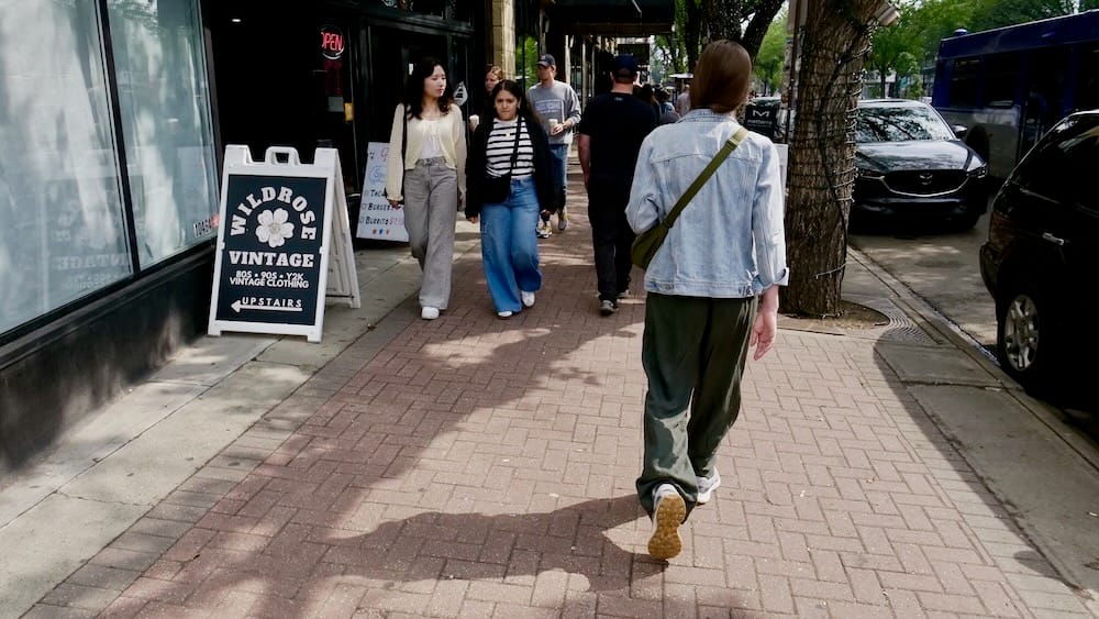 Whyte Avenue, Old Strathcona, Edmonton, Alberta, Canadá Audrey Bergner caminando por una vereda de ladrillos transitada en Whyte Avenue pasando el cartel de la tienda de ropa Wildrose Vintage, con peatones y autos estacionados en Old Strathcona, Edmonton, Alberta, Canadá