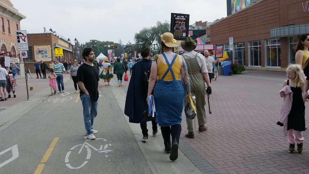 Street scene during the Edmonton Fringe Festival, Old Strathcona, Alberta, Canada Costumed festivalgoers and a clown walk down a bike-lane-marked street lined with brick buildings and show posters during the Edmonton Fringe Festival in Old Strathcona, Edmonton, Alberta, Canada.