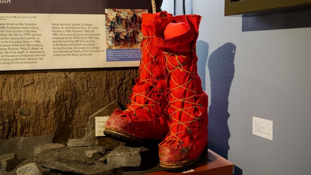 Pair of bright red vintage mountaineering boots displayed on rocks inside the Whyte Museum in Banff, Alberta, Canada, beside interpretive panels about climbing history in the Rocky Mountains.