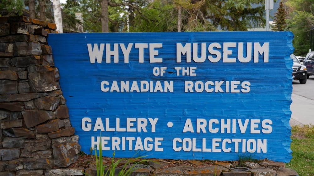 Blue exterior sign for the Whyte Museum of the Canadian Rockies in Banff, Alberta, Canada, identifying its gallery, archives and heritage collection beside a stone wall and trees.