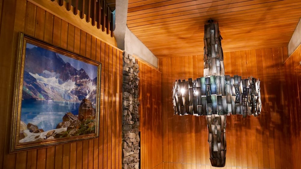Mountain lake landscape painting, Whyte Museum of the Canadian Rockies, Banff, Alberta A framed mountain and glacial lake landscape painting hangs on a wood-paneled wall beside a large stained-glass cross-shaped light fixture in a hallway at the Whyte Museum of the Canadian Rockies, Banff, Alberta, Canada.