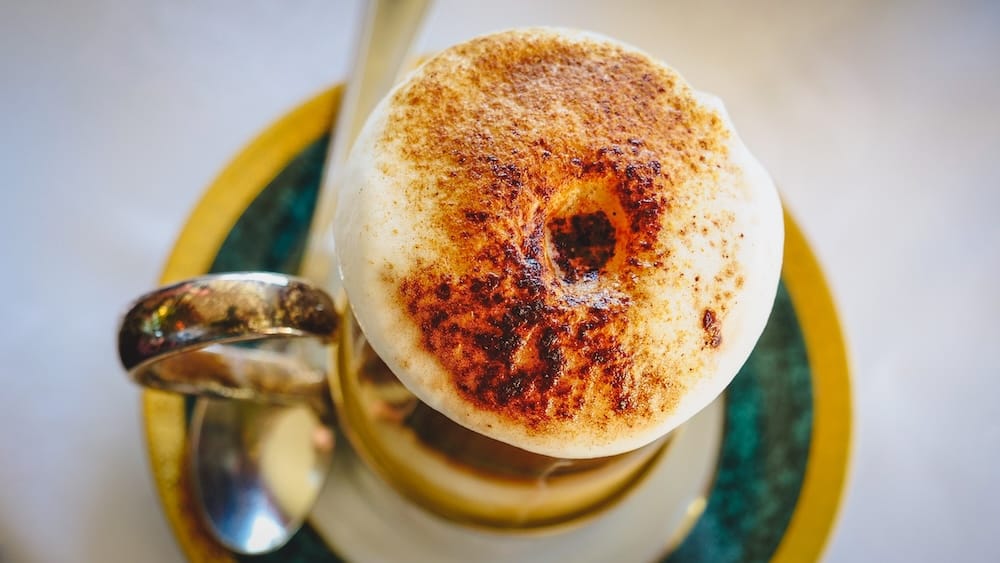 Cappuccino with Toasted Foam During Afternoon Tea at Llao Llao Hotel, Bariloche, Patagonia, Argentina Overhead close-up of a cappuccino with thick toasted foam served during afternoon tea at Llao Llao Hotel in Bariloche, Río Negro, Patagonia, Argentina, presented on a decorative porcelain cup and saucer with a silver spoon.