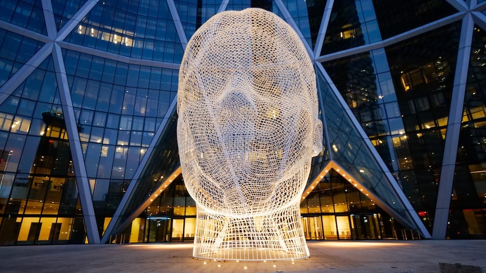 Wonderland Sculpture Downtown Calgary Alberta Canada Public Art Installation Beside Modern Glass Architecture Wonderland sculpture in downtown Calgary, Alberta, Canada, showing the large illuminated wire-mesh human head framed by the glass and steel facade of a modern office building.