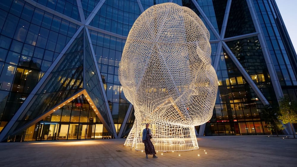 Audrey Bergner en la escultura Wonderland junto a The Bow, centro de Calgary, Alberta, Canadá Audrey Bergner está bajo la escultura iluminada Wonderland con forma de cabeza de alambre, frente a The Bow en el centro de Calgary, Alberta, Canadá, con la fachada geométrica de vidrio de la torre detrás.