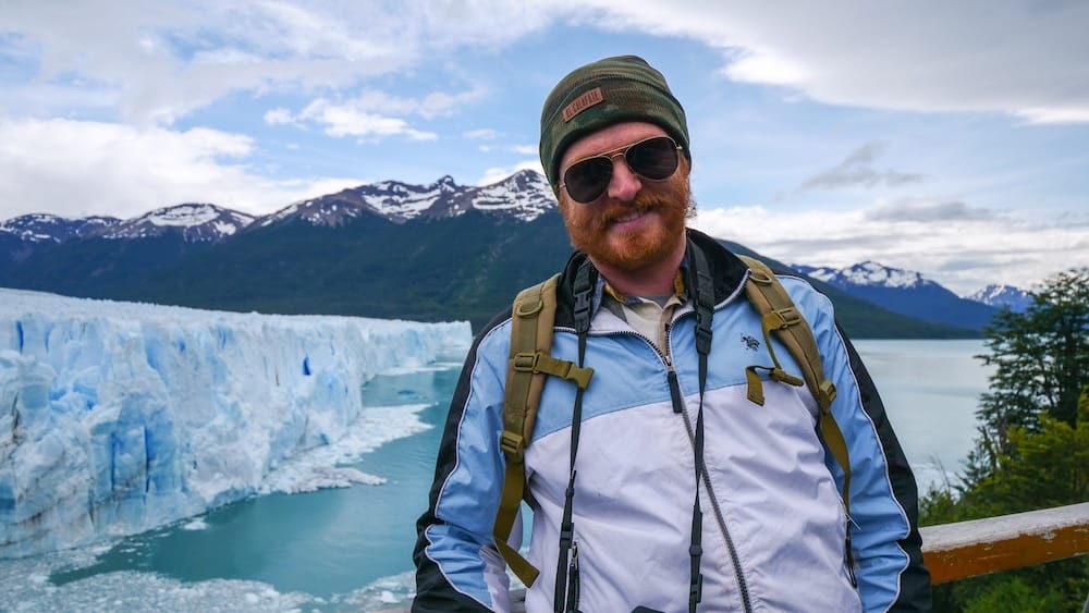 Boardwalk viewing platform, Perito Moreno Glacier, El Calafate, Santa Cruz, Argentine Patagonia — Samuel Jeffery Samuel Jeffery smiling on a wooden boardwalk railing with the towering blue ice wall of Perito Moreno Glacier and snow-capped Andes behind him, El Calafate, Santa Cruz, Argentine Patagonia.