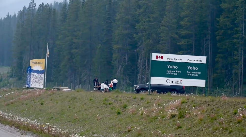Yoho National Park and British Columbia welcome signs beside the highway in Canada Parks Canada’s Yoho National Park sign stands beside a Welcome to British Columbia sign along the roadside, with evergreen forest, parked vehicles and a grassy verge surrounding the park entrance area.