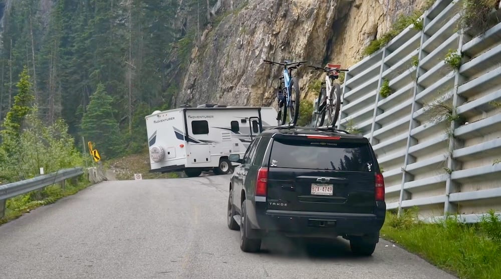 A vehicle towing a travel trailer with mountain bikes follows an SUV along a narrow switchback section of Yoho Valley Road toward Takakkaw Falls in Yoho National Park, British Columbia, with a rock-cut avalanche barrier lining the road.