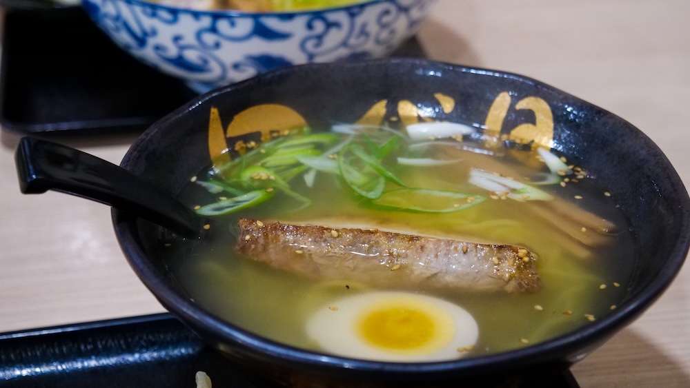 Primer plano de shio ramen en Yondaime Ramen en Hakodate, Hokkaido, Japón, servido en un bol negro con caldo dorado claro a la sal, cerdo asado, medio huevo duro, cebolla de verdeo, brotes de bambú y fideos.