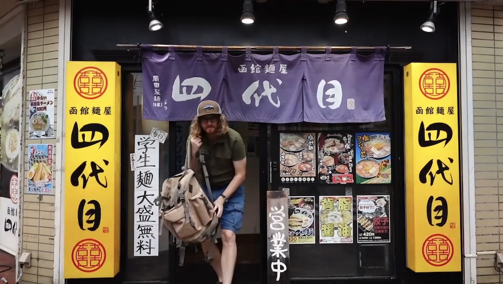 Samuel Jeffery with a backpack steps out under purple entrance curtains reading Hakodate Menya Yondaime, between tall yellow signs and posters of ramen bowls, at a ramen shop in Hakodate, Hokkaido, Japan.