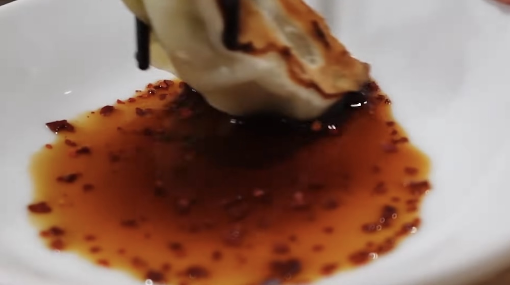 Close-up of a browned gyoza held in black chopsticks and dipped into a white dish of dark soy sauce with red chili flakes at a ramen shop in Hakodate, Hokkaido, Japan.