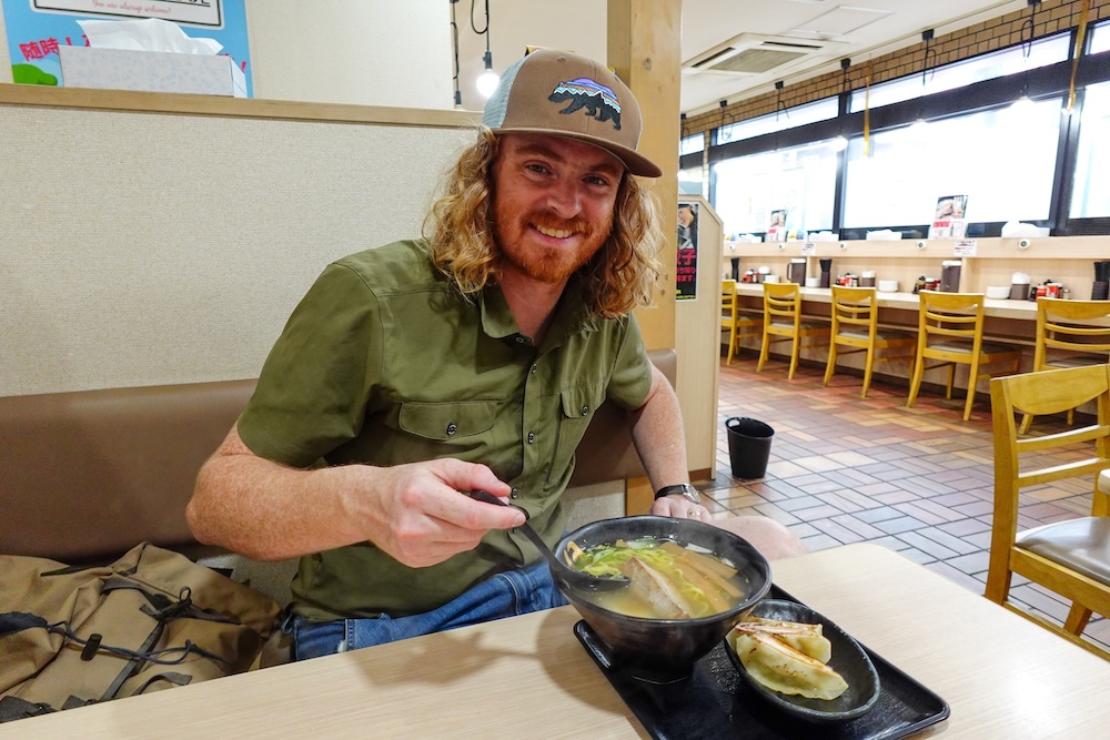 Samuel Jeffery in a green shirt and brown cap smiles over a black bowl of salt ramen with sliced pork and a small plate of gyoza at a booth table in a ramen shop in Hakodate, Hokkaido, Japan.