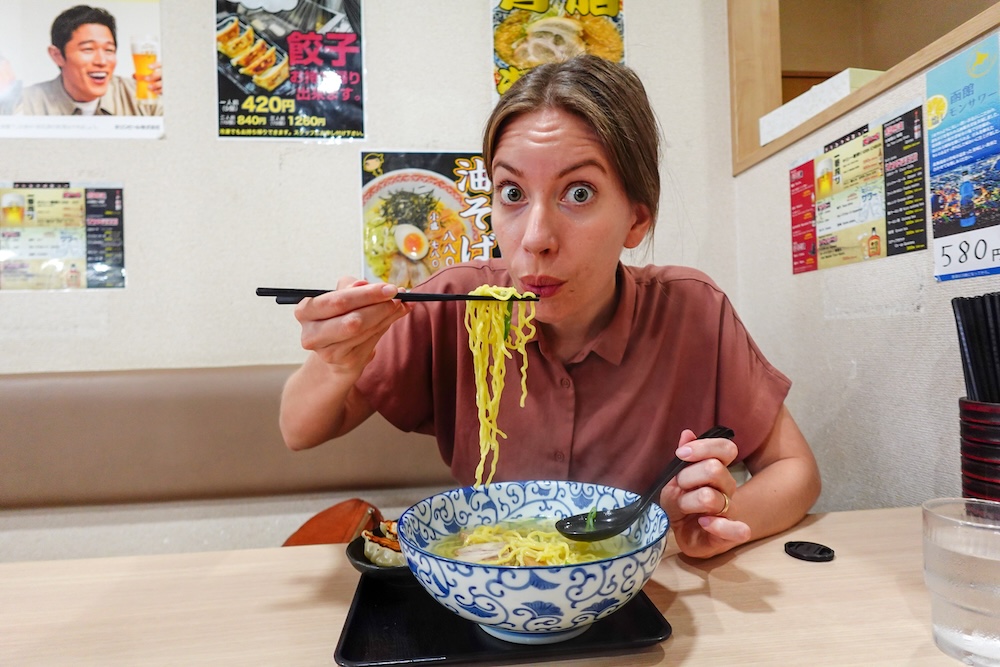 Audrey Bergner eating noodles from her salt ramen at the Yondaime ramen shop, Hakodate, Hokkaido, Japan Audrey Bergner lifts yellow, slightly wavy noodles on chopsticks above a blue-and-white bowl of salt ramen at a booth in the Yondaime ramen shop in Hakodate, Hokkaido, Japan, with gyoza and ramen posters behind her.