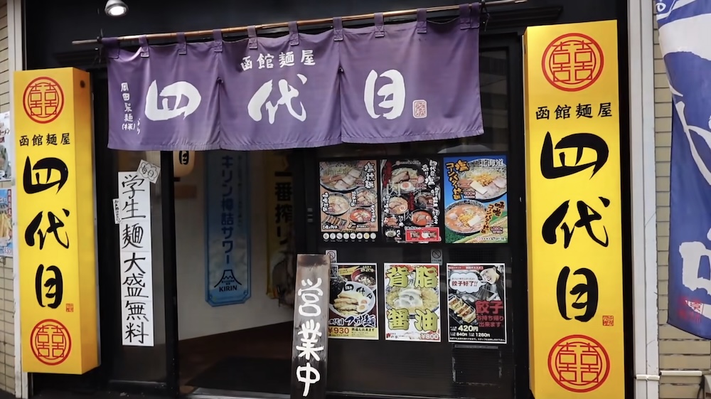 Yondaime ramen shop storefront entrance with purple noren and yellow signs in Hakodate, Hokkaido, Japan The storefront entrance of the Yondaime ramen shop in Hakodate, Hokkaido, Japan, features a purple noren curtain with white kanji, matching vertical yellow pillar signs on either side, and illustrated ramen and gyoza menu posters on the glass.