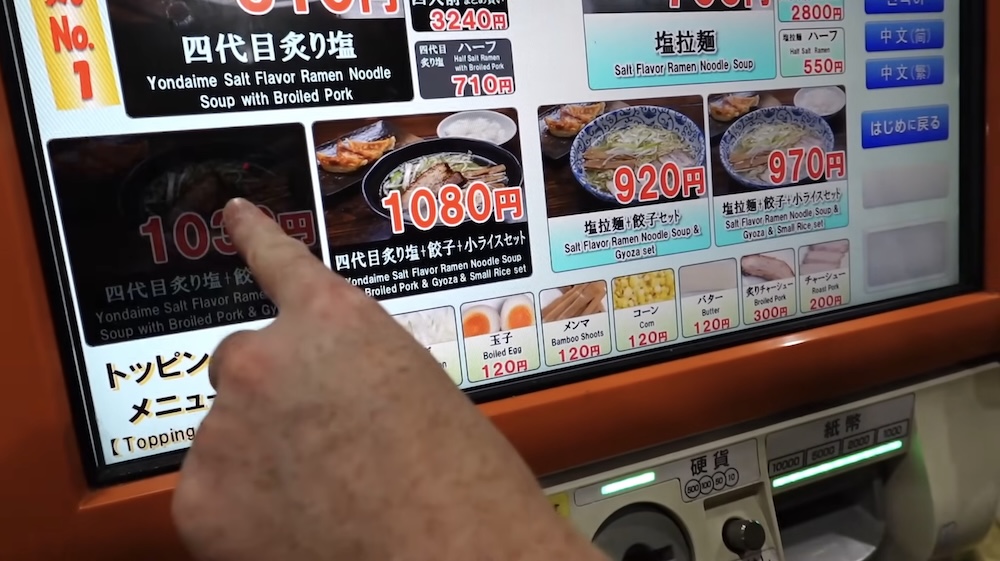 A finger points at the seared salt ramen with broiled pork and gyoza tile on the touchscreen of a ramen ticket machine in Hakodate, Hokkaido, Japan, with half-size bowls listed at the top right and a toppings row below.