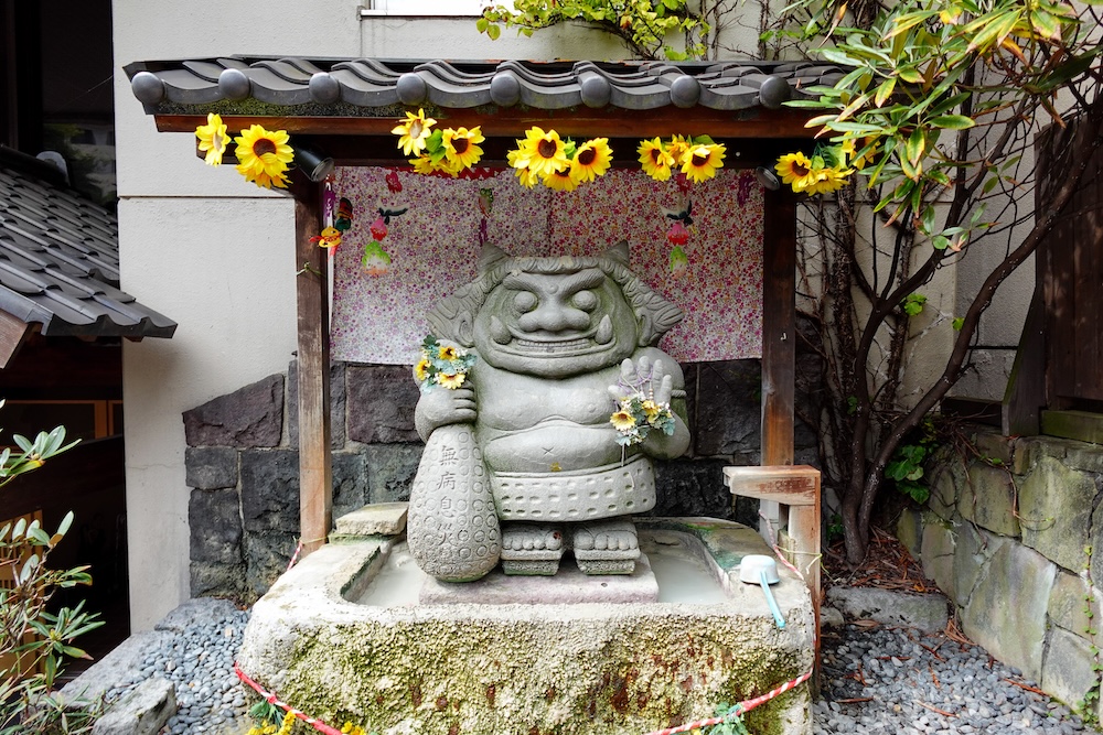 Estatua gris de piedra tallada del demonio de la salud Yukake Kizo, de pie en una pileta de agua lechosa con un cucharón, un garrote kanabo y girasoles amarillos bajo el techo de tejas de un santuario en Noboribetsu Onsen, Hokkaido, Japón.