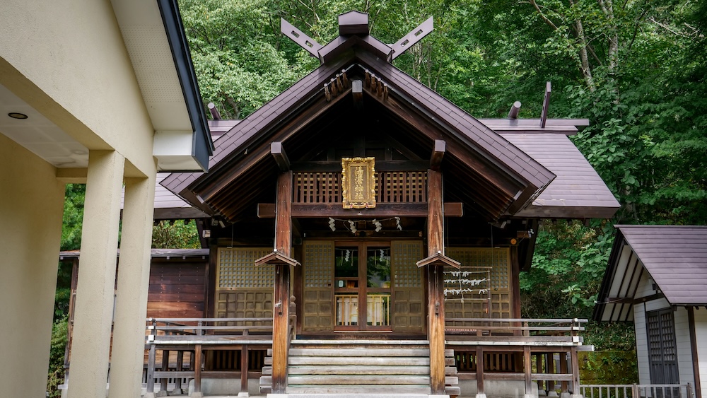 Salón principal del santuario Yuzawa y su arquitectura sintoísta de madera en el bosque sobre Noboribetsu Onsen, Hokkaido, Japón El salón principal de madera del santuario Yuzawa, con remates cruzados en el techo, una placa dorada con el nombre y tiras de papel blanco, rodeado de árboles verdes en Noboribetsu Onsen, Hokkaido, Japón.