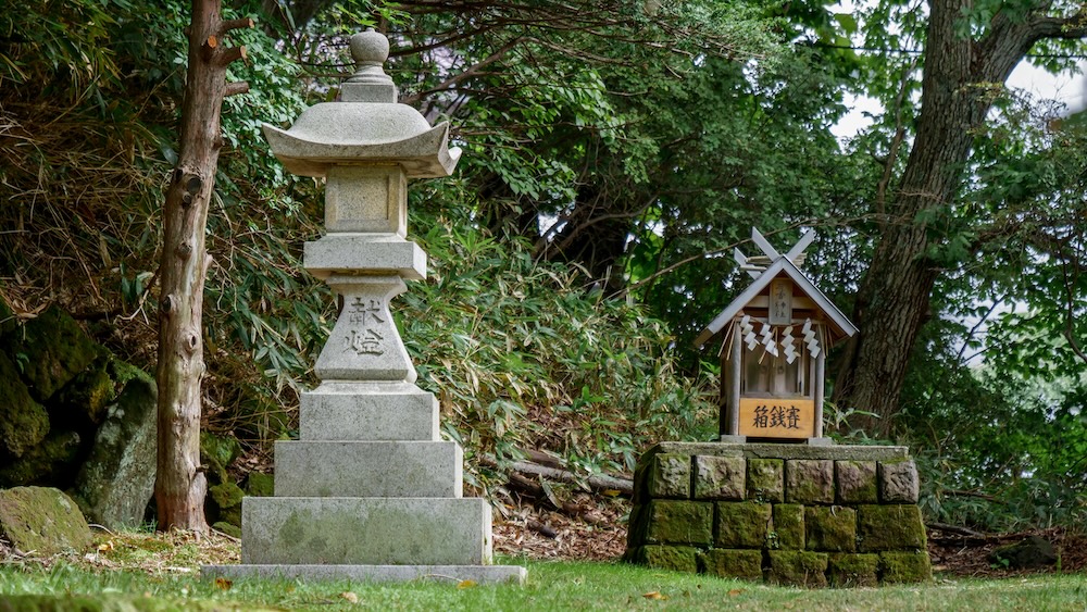 Farol tradicional de piedra junto a una pequeña estructura de santuario sintoísta de madera decorada con tiras de papel blanco en Yuzawa Shrine, Noboribetsu Onsen, Hokkaido, Japón, rodeada de piedras con musgo, bambú bajo y vegetación boscosa.