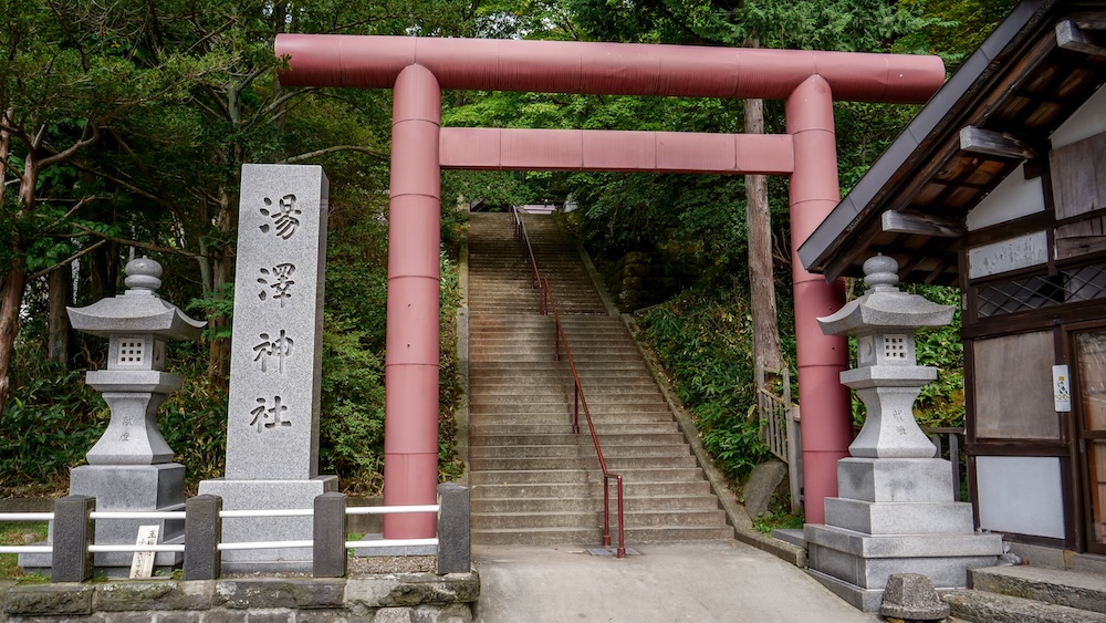 Portal torii rojo y escalera de piedra de entrada al santuario Yuzawa en Noboribetsu Onsen, Hokkaido, Japón Un portal torii rojo enmarca la larga y empinada escalera de piedra que sube al santuario Yuzawa, flanqueada por faroles de piedra y un pilar tallado entre los árboles en Noboribetsu Onsen, Hokkaido, Japón.